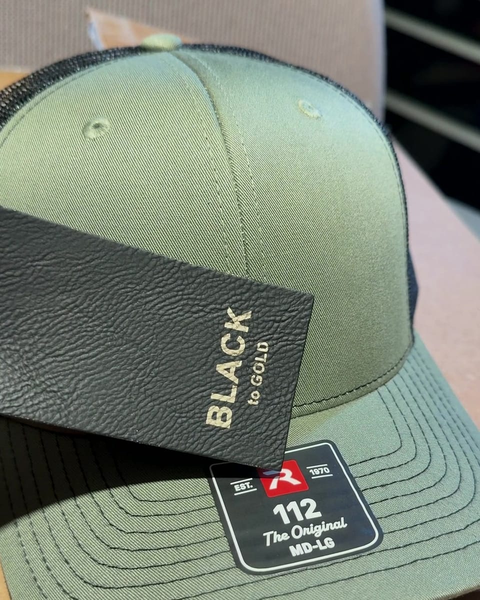 A piece of black leatherette, labeled Black to gold, held against a Loden/Black hat