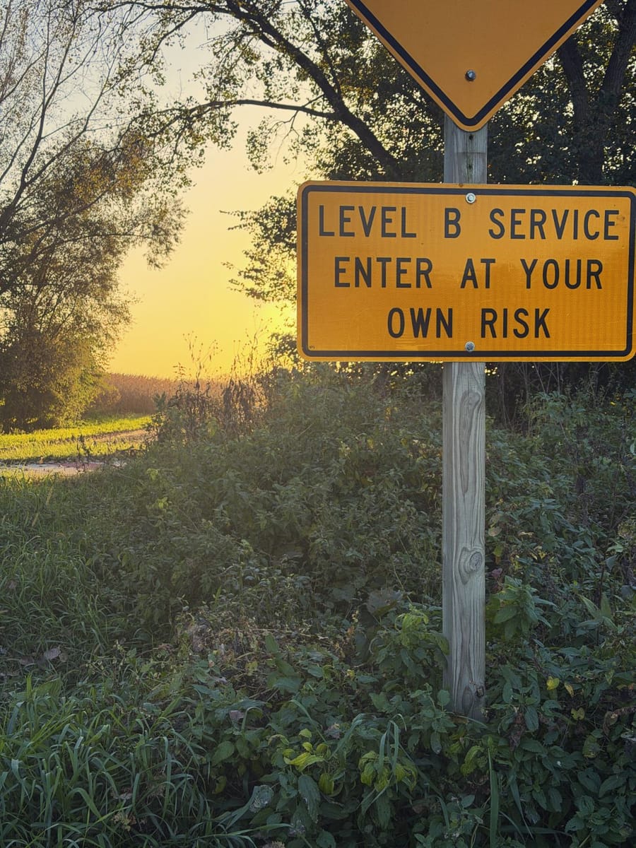 A yellow Level B Service, Enter at your own risk sign on a wooden post beside a gravel road at sunset